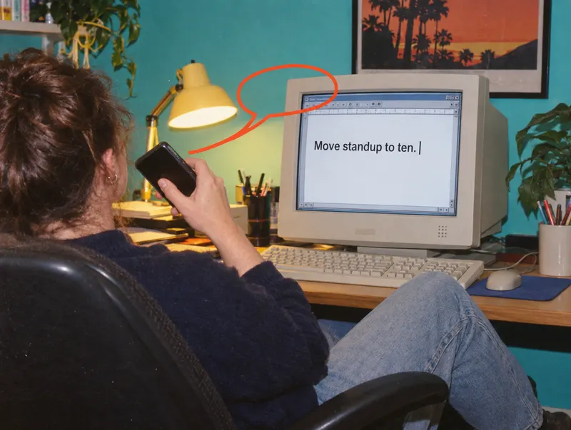 A person in a desk chair talks into a phone while their words, Move standup to ten., appear on a beige computer screen.